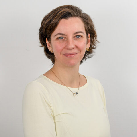 Eine Person mit kurzen, dunklen Haaren und einem hellen Oberteil steht vor einem schlichten Hintergrund. Sie lächelt leicht und trägt kleine, runde Ohrringe und eine zarte Halskette.