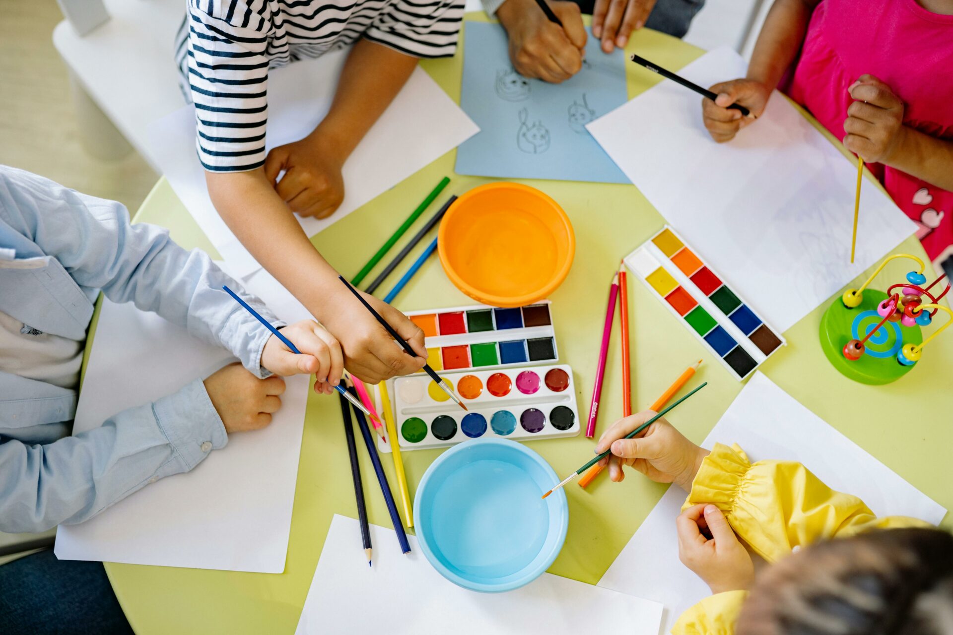 Kinder sitzen um einen Tisch und zeichnen und malen mit Wasserfarben, Buntstiften und Papier. Man kann ihre Hände sehen, während sie Kunstwerke schaffen. Auf der gelben Tischplatte stehen Malutensilien und eine Schale mit Wasser.